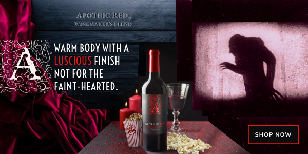 A dark interior space with a table, movie screen, and red velvet curtain. A wine bottle, wine glass, candles and box of popcorn are on the table. The silhouette of a vampire appears on the movie screen.