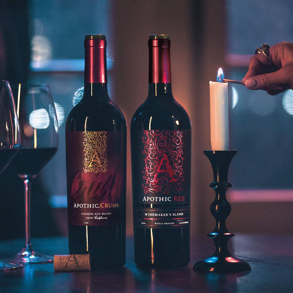 A table with two wine bottles, a candle stick, and a filled wine glass. A hand reaching in from the left lights the candle.