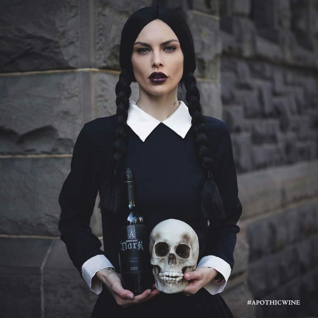 A young white woman with long dark braids holding a skull and a wine bottle.