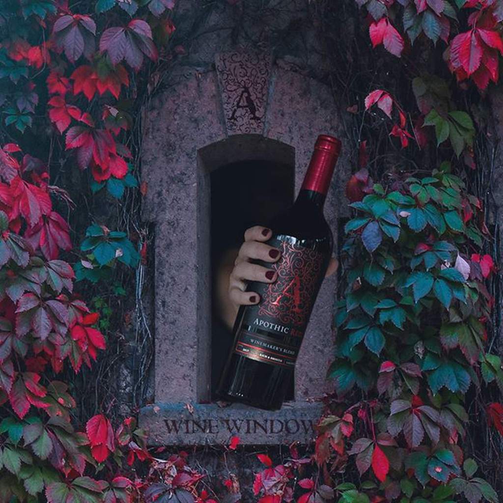 A tomb in the ground with a small opening surrounded by foliage. A hand holding a wine bottle reaches out of the opening.