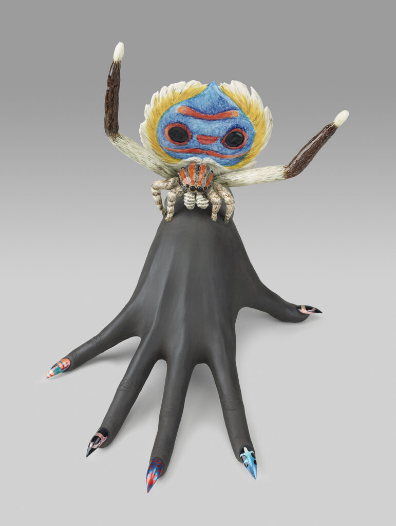 A small colourful figure sitting on top of a dark grey ceramic hand with long colourful fingernails. 