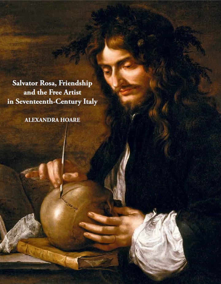 A young white man with long brown hair, moustache and beard looking down on a skull in his hands and writing on the skull with a quill.