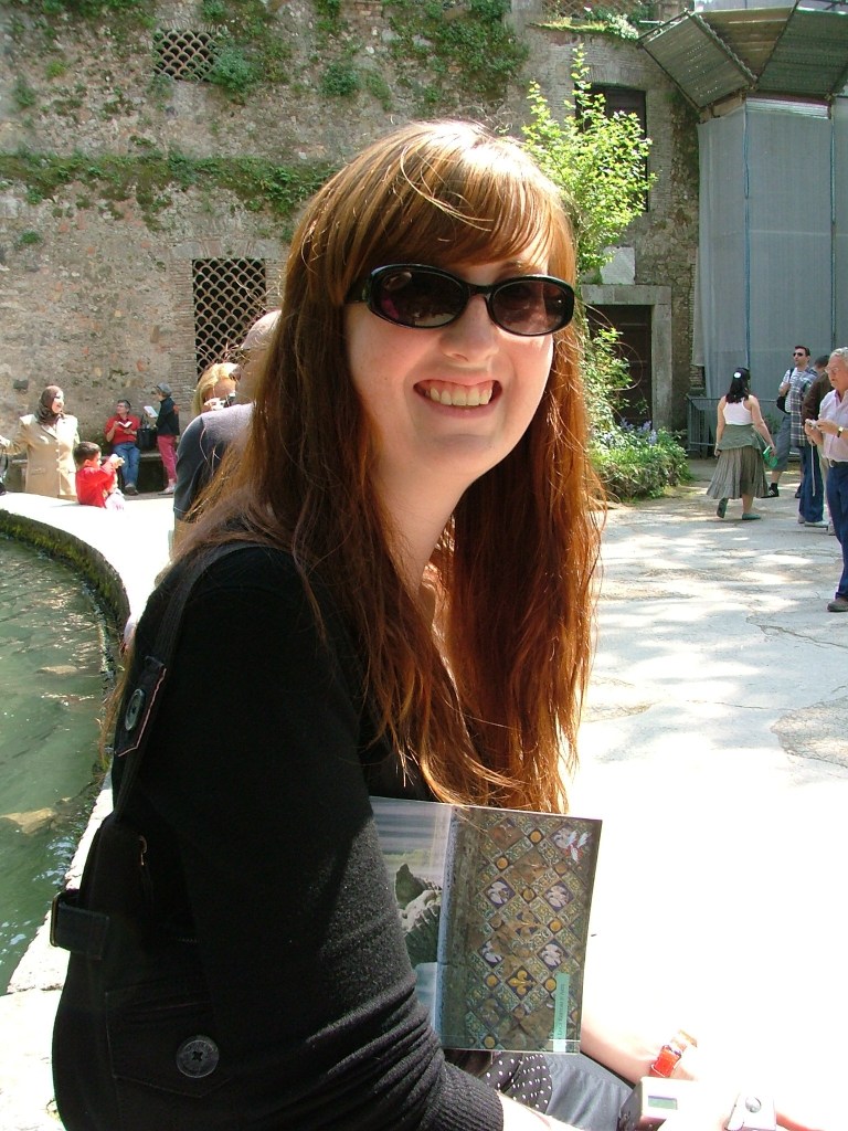 A hip-length photo of a smiling white woman with long red hair and sunglasses sitting outside on the edge of a fountain.